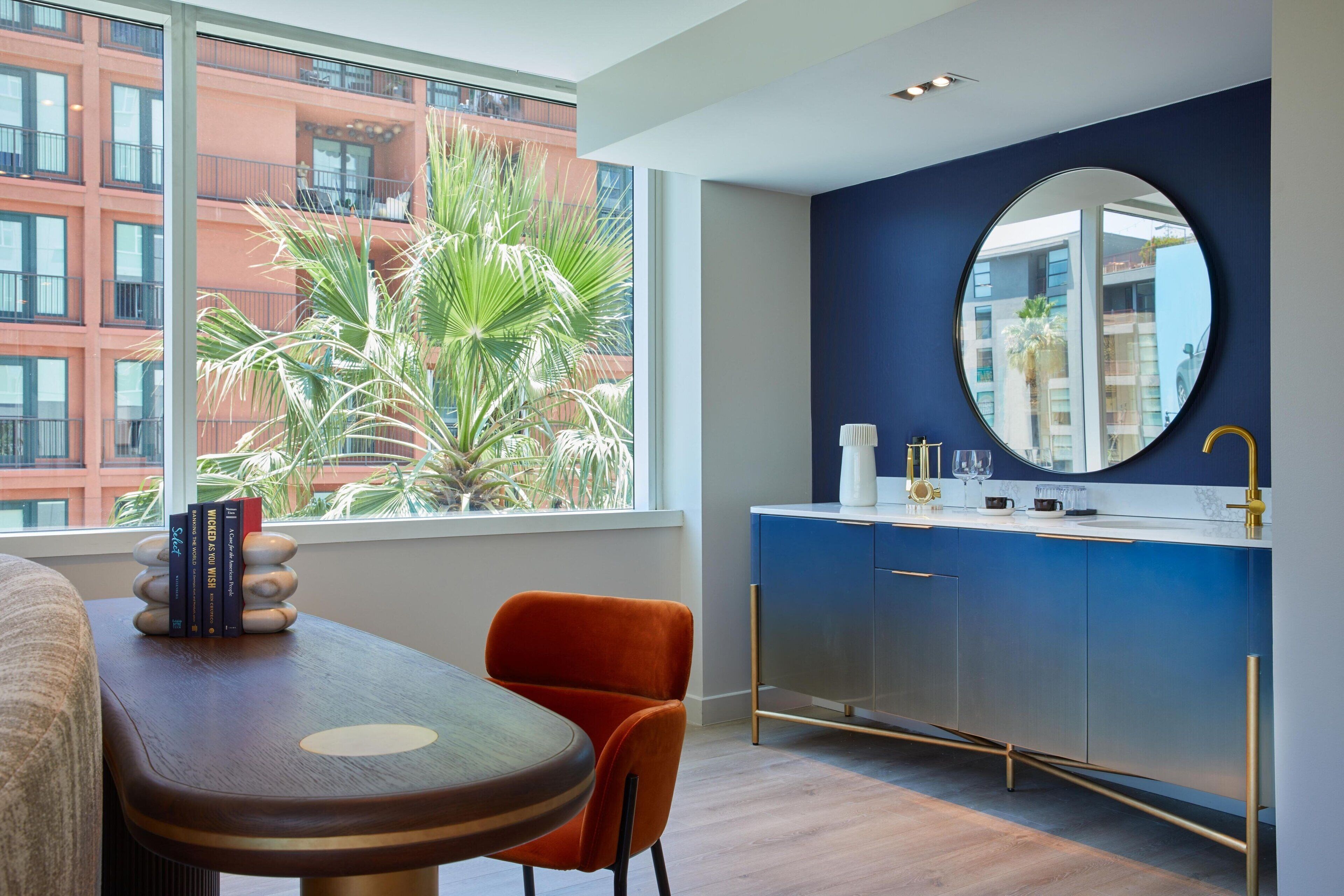 Ein modernes Hotelzimmer mit großem Fenster, einem runden Spiegel, einem Holztisch, einem orangefarbenen Sessel und einem blauen Schrank mit goldener Armatur.