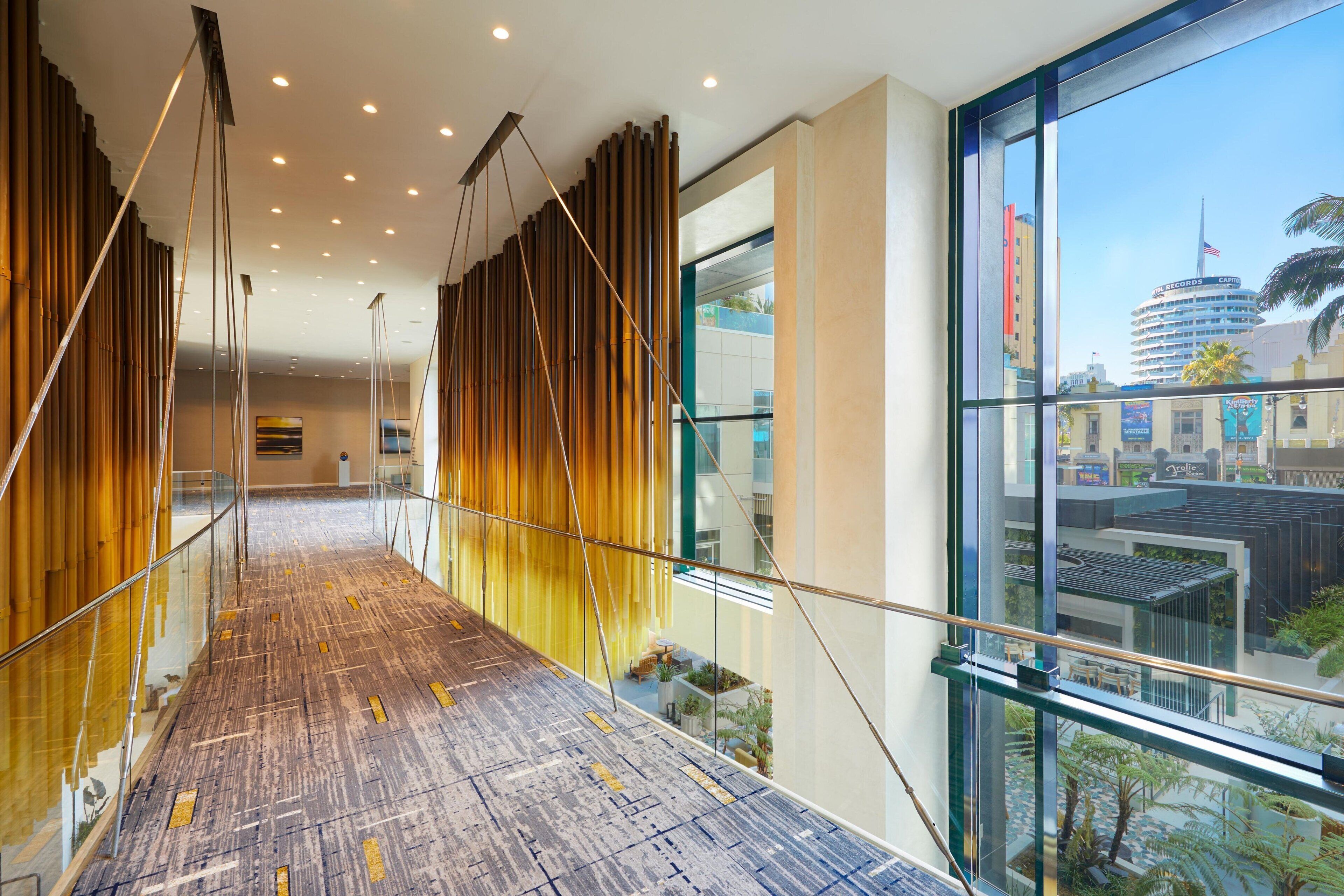Un couloir d’hôtel moderne avec des rampes de protection en verre, un plancher en bois et une vue sur le paysage urbain grâce à de grandes fenêtres.