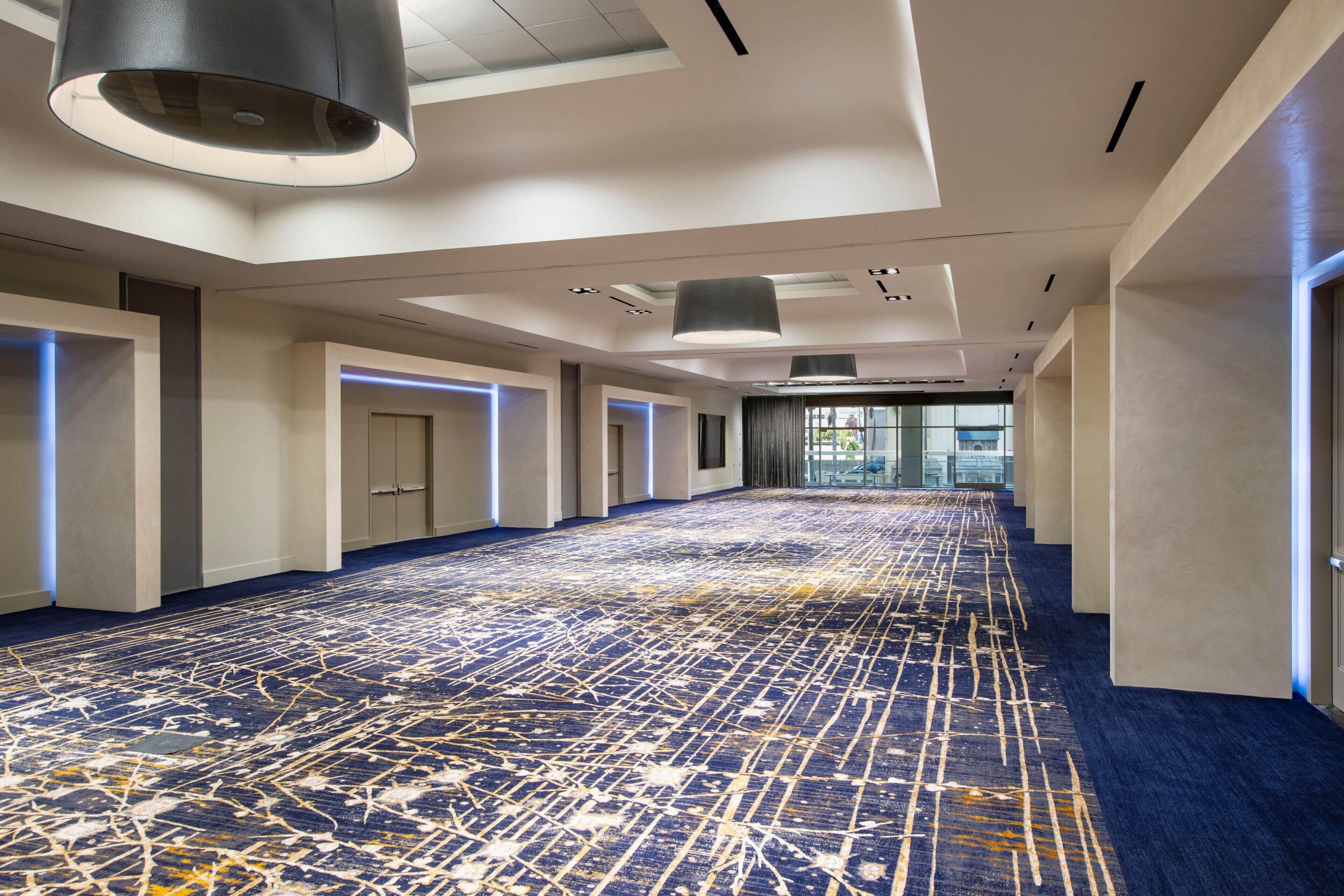 A spacious hotel corridor with a patterned carpet, recessed lighting, and a view of the outdoors through large windows.