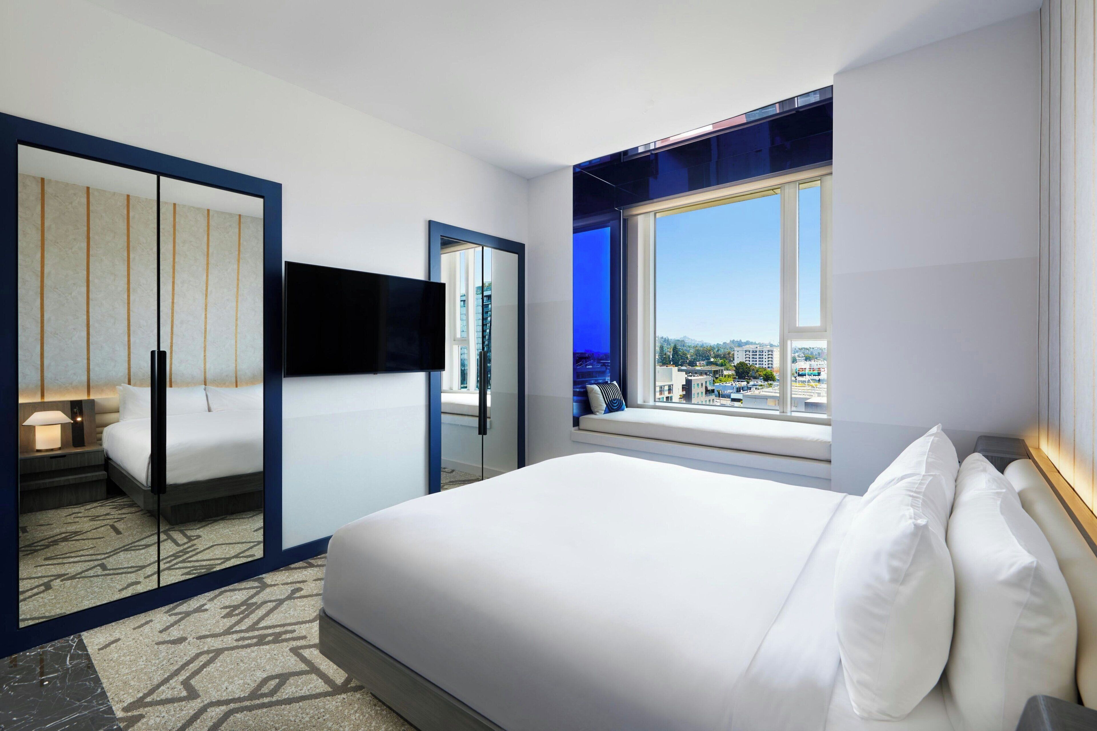 A hotel room with a large bed, a TV, a window with a city view, and a sliding door with a mirror.