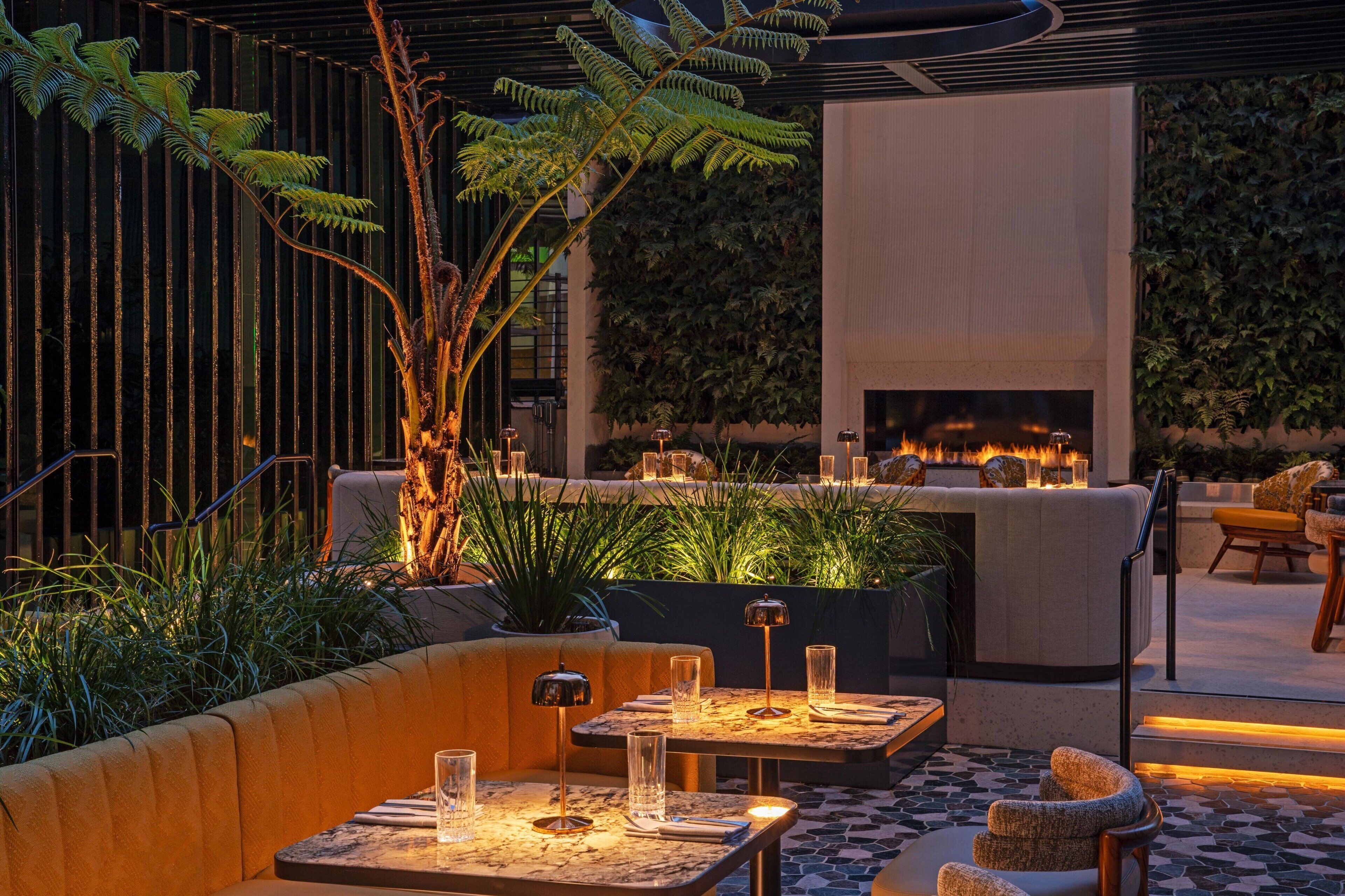 An outdoor seating area with tables and chairs, surrounded by plants and a fire feature.