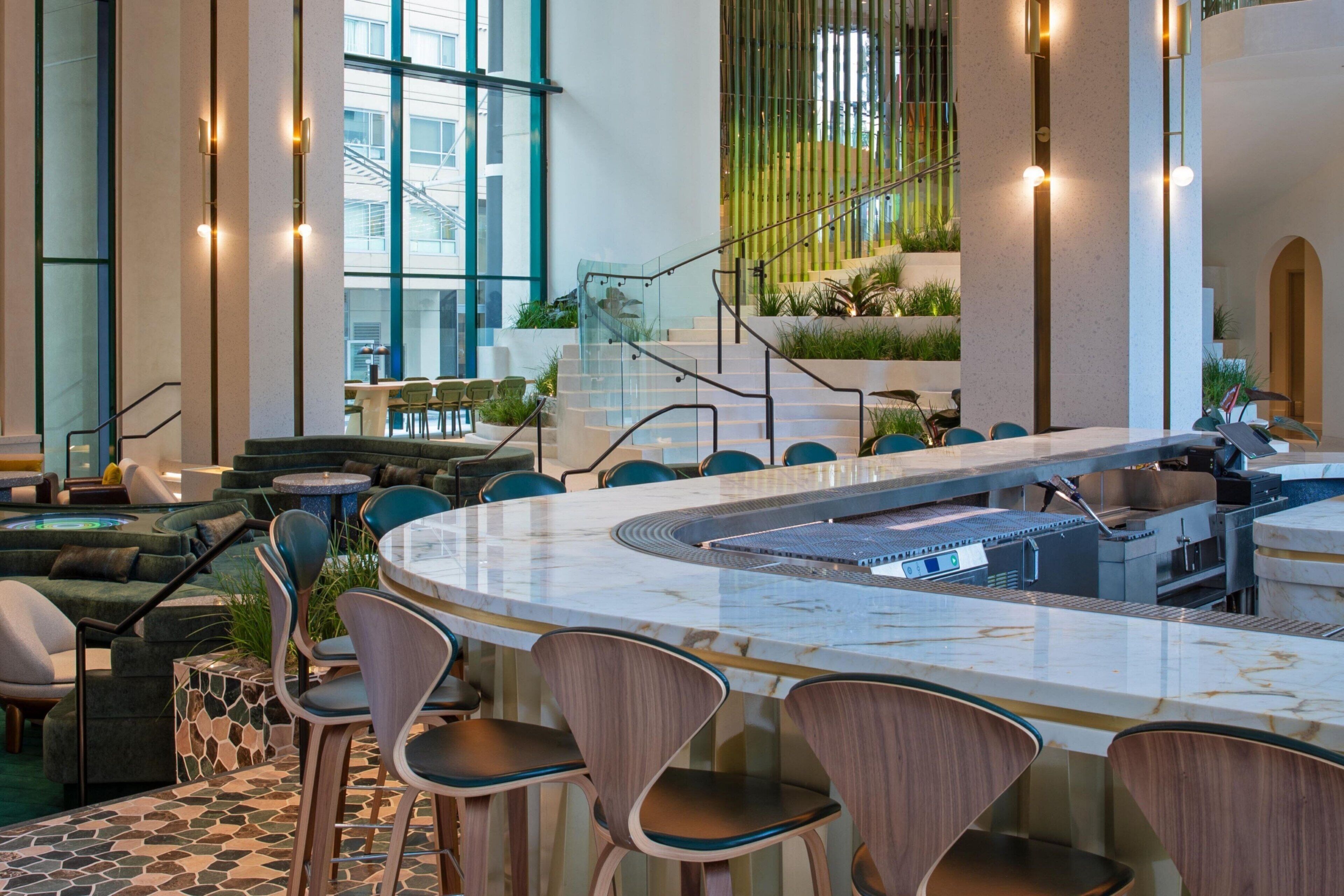 A modern indoor space with a central bar area, seating arrangements, and a staircase leading to an upper level.