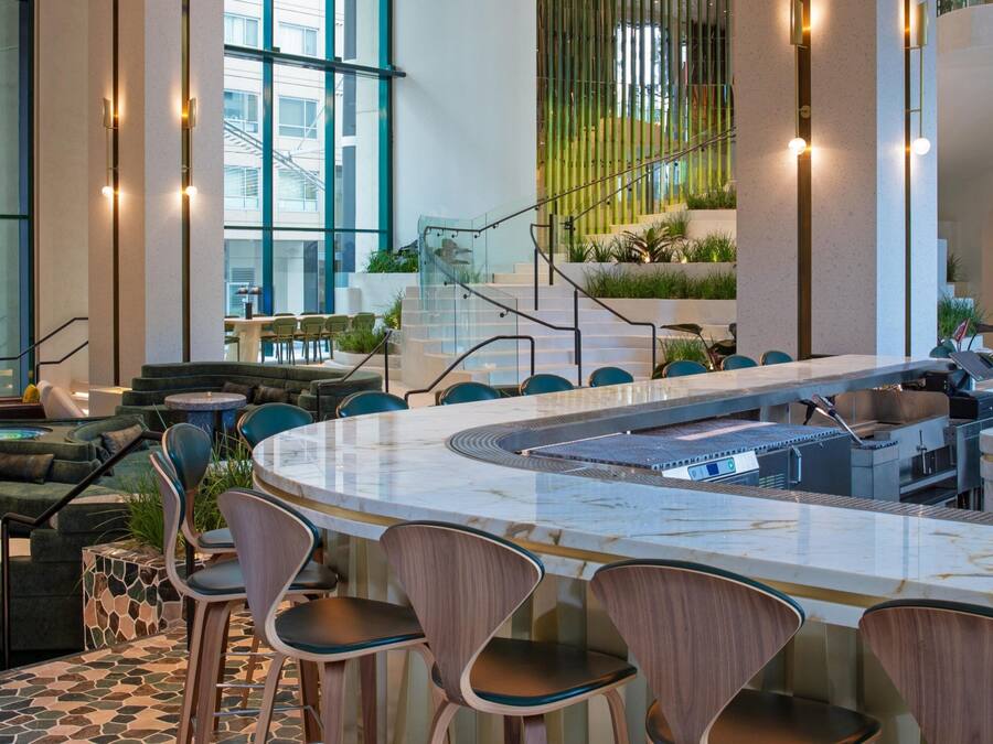 A modern indoor space with a central bar area, seating arrangements, and a staircase leading to an upper level.