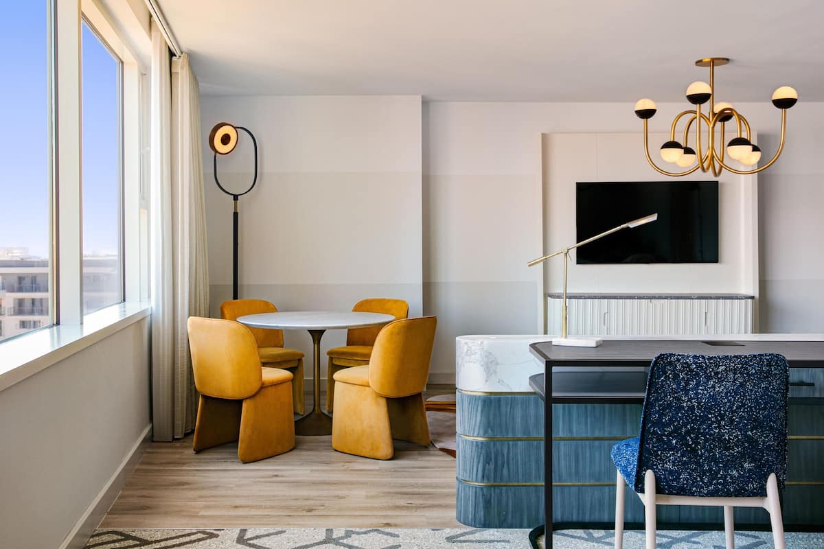 A modern hotel room with a large window, a round dining table with yellow chairs, a blue accent chair, a white sofa, and a chandelier.