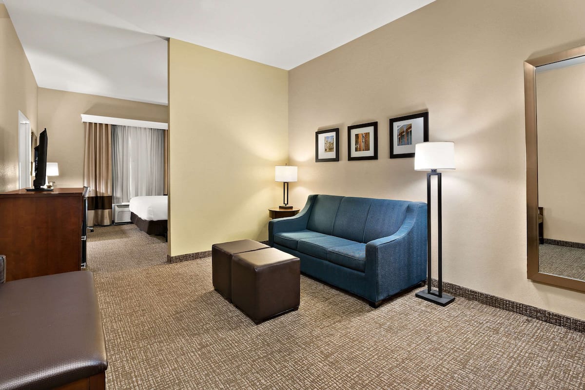 Habitación de hotel con un sofá azul, un pequeño reposapiés, un escritorio con lámpara, una cama y un espejo grande.