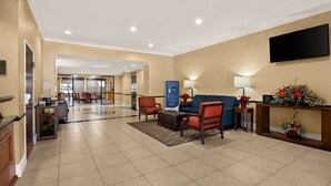 Un vestíbulo de hotel con mostrador de recepción, zona de estar con sillas, televisor montado en la pared y una planta decorativa.