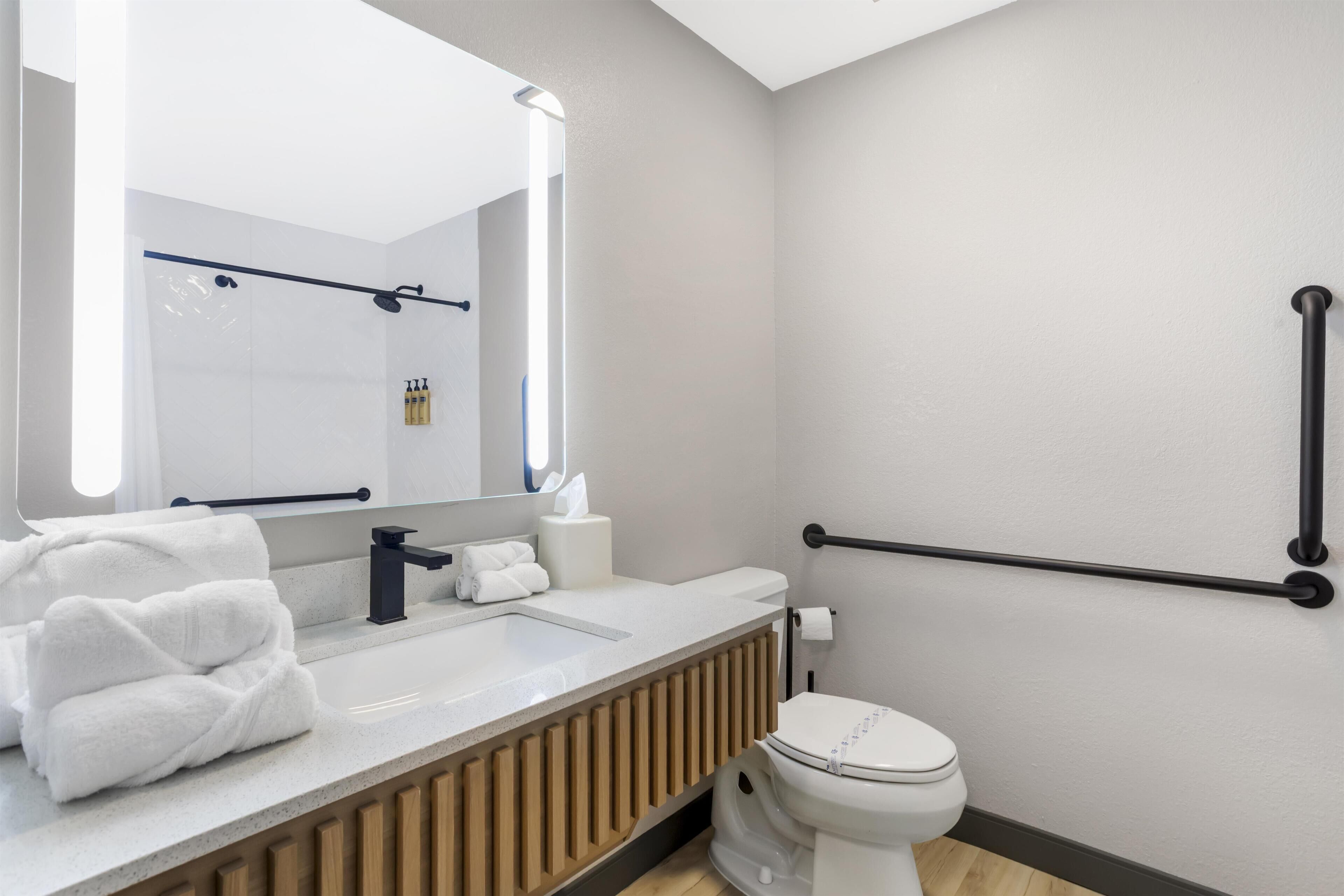 A modern bathroom with a white sink, black faucet, and a toilet. There are towels on the sink and a handrail on the wall.