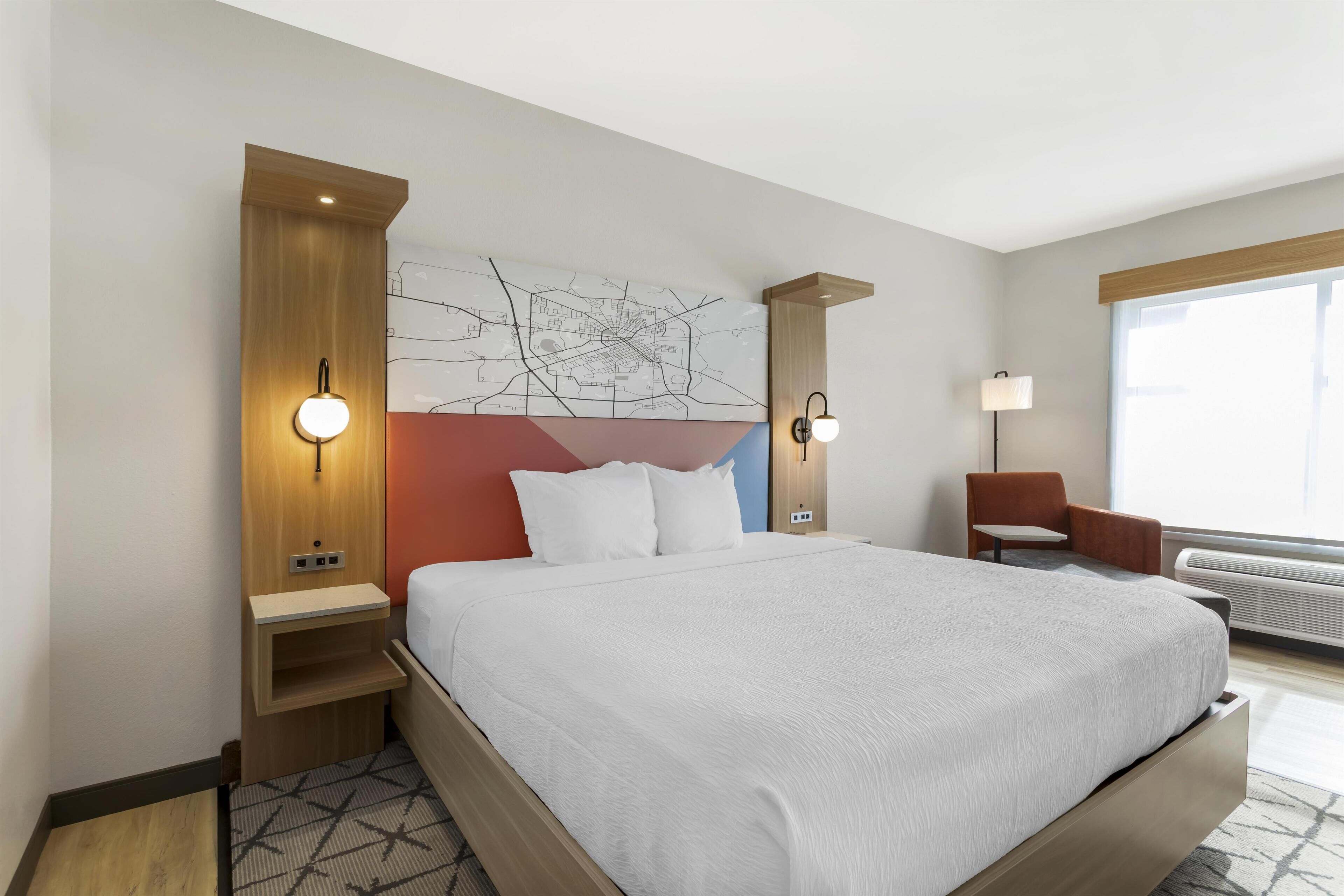 Una habitación de hotel moderna con una cama grande, un mapa colgado en la pared, una mesita de noche, una silla y una ventana con luz natural.