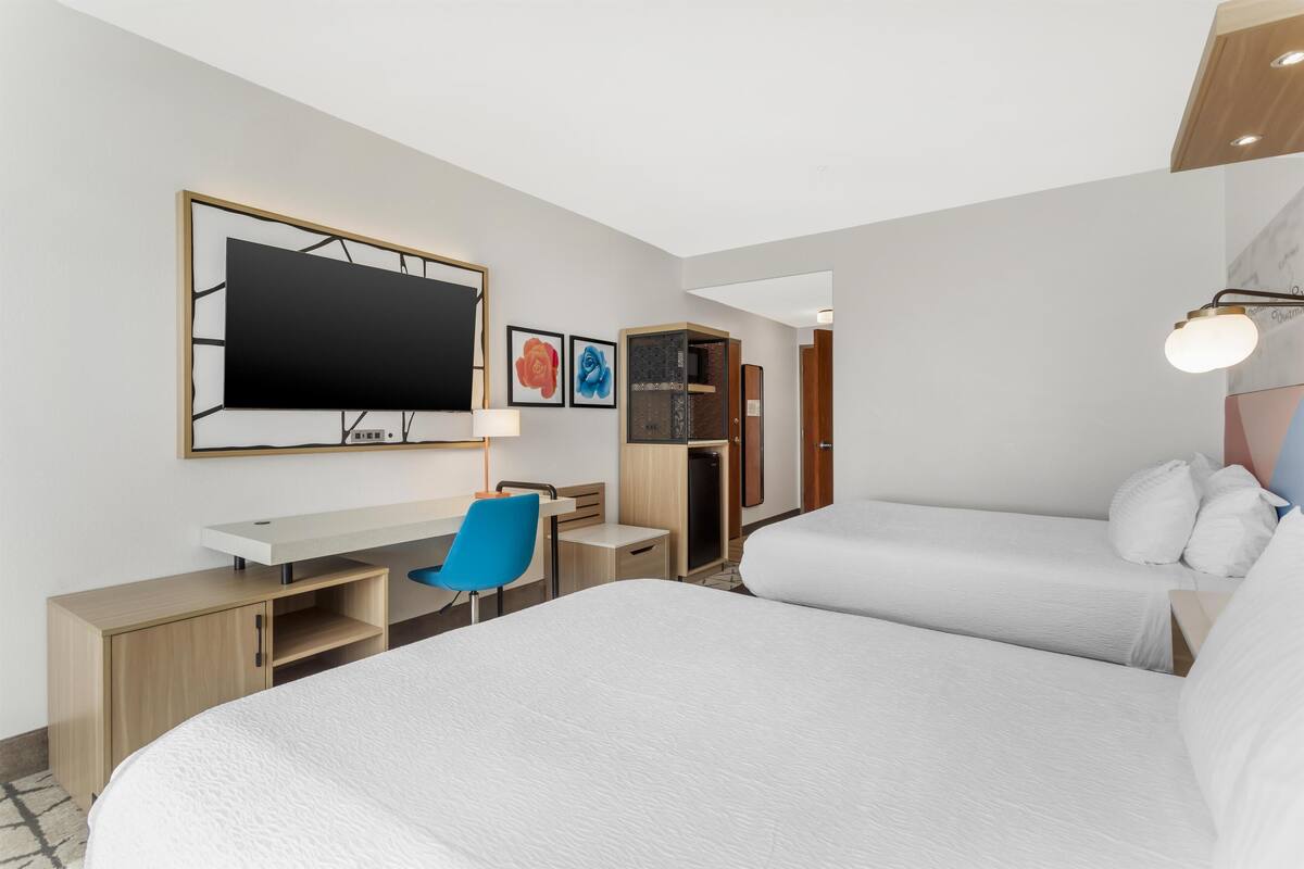 A hotel room with two beds, a desk, a chair, a TV, and artwork on the wall.