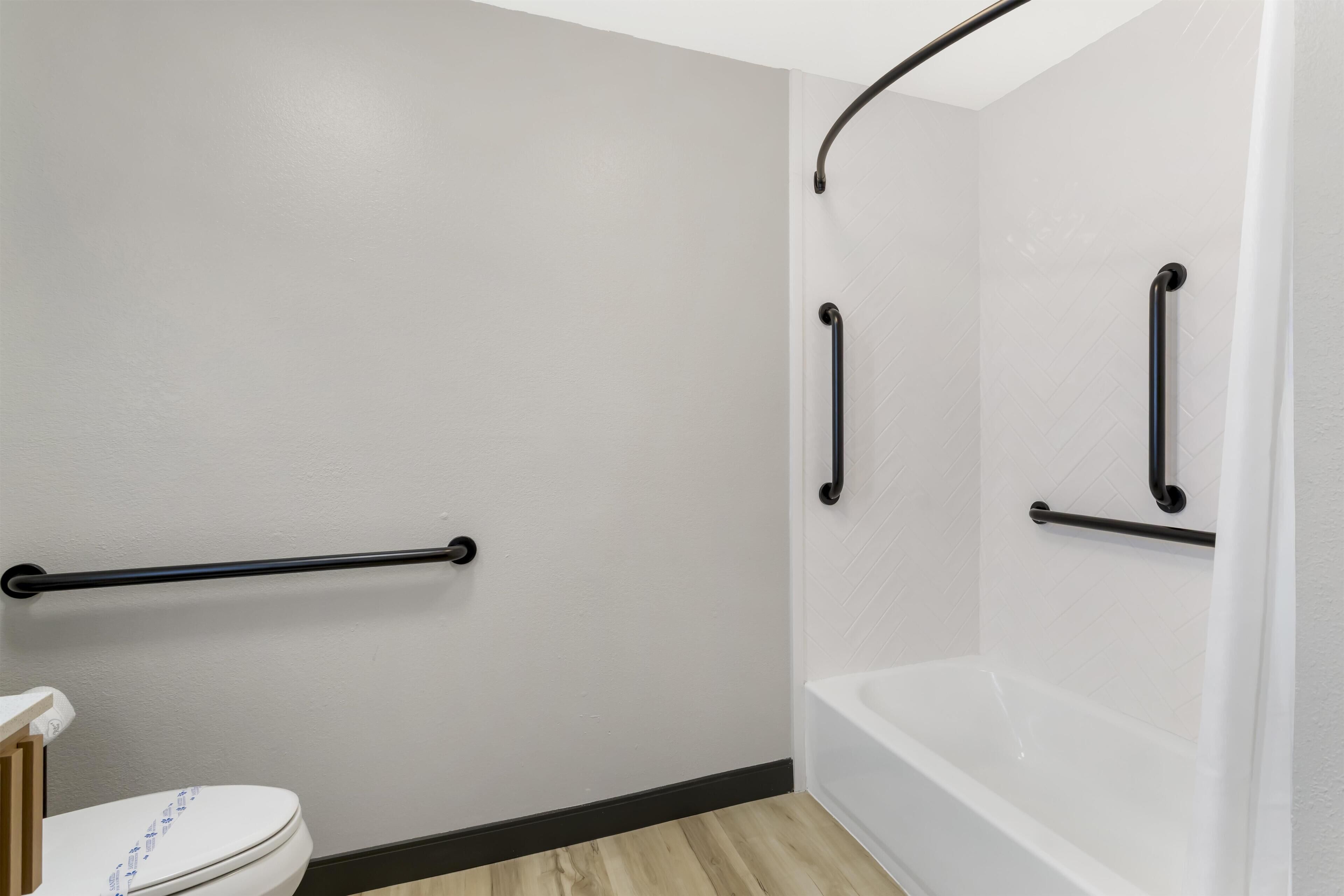 Un baño con inodoro blanco, una bañera con una barra de apoyo negra y una zona de ducha con una barra de apoyo negra curva.