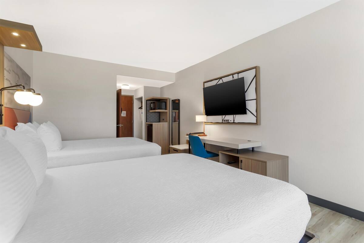A hotel room with two beds, a desk, a TV, and a kitchenette.