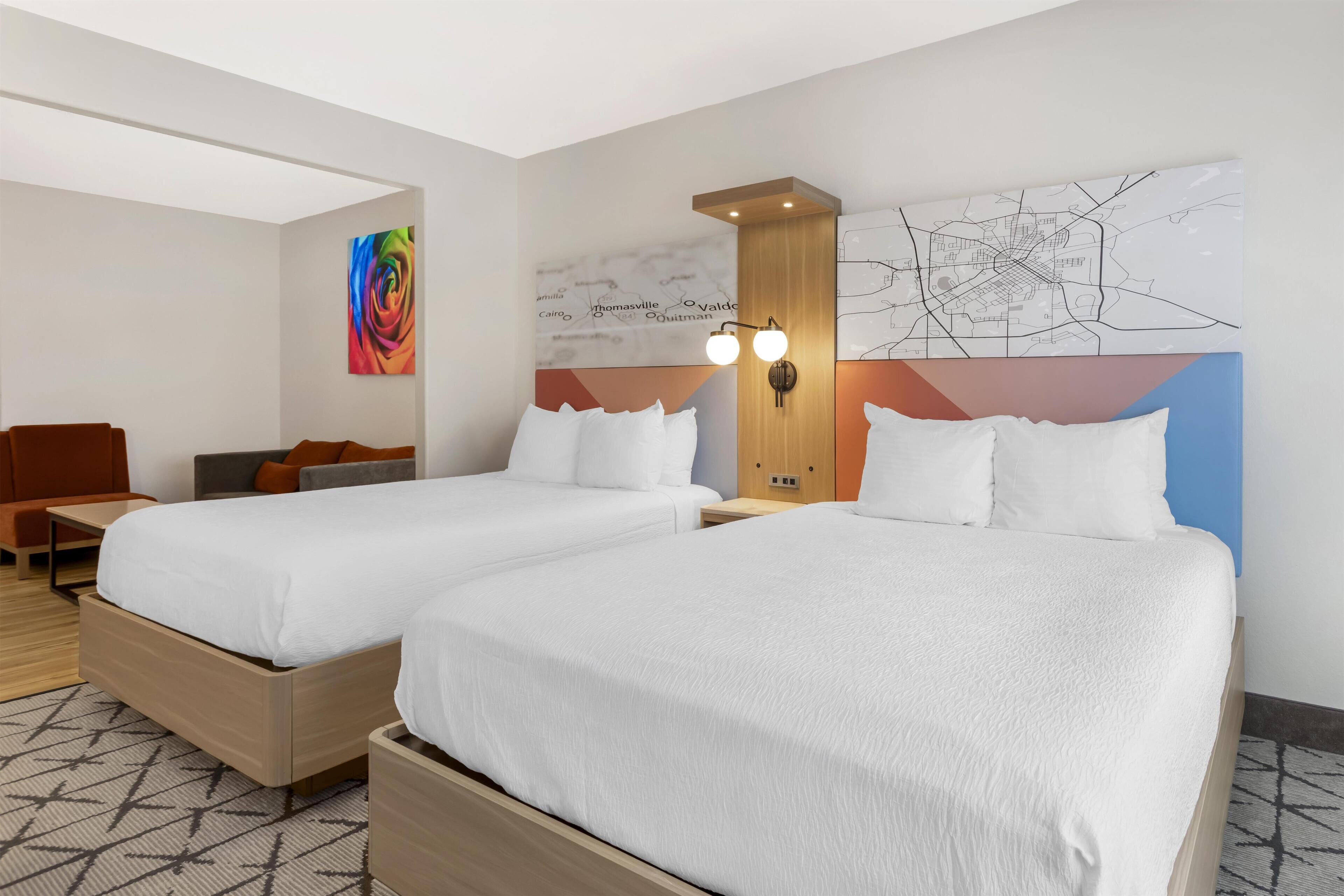Habitación de hotel con dos camas, un mural de mapa, un cuadro abstracto colorido y una pequeña zona de estar con una mesa y sillas.
