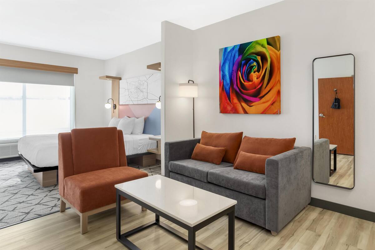 A modern hotel room with a bed, a grey sofa, an orange armchair, a white table, a colorful abstract painting, a floor lamp, and a mirror on the wall.