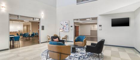Un hall d’entrée spacieux avec un plafond haut, un espace de détente central agrémenté de canapés et de fauteuils, un téléviseur à écran plat fixé au mur et un escalier menant à un niveau supérieur.
