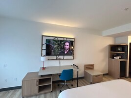 A modern hotel room with a wooden desk, a blue chair, a bed, and a TV mounted on the wall.