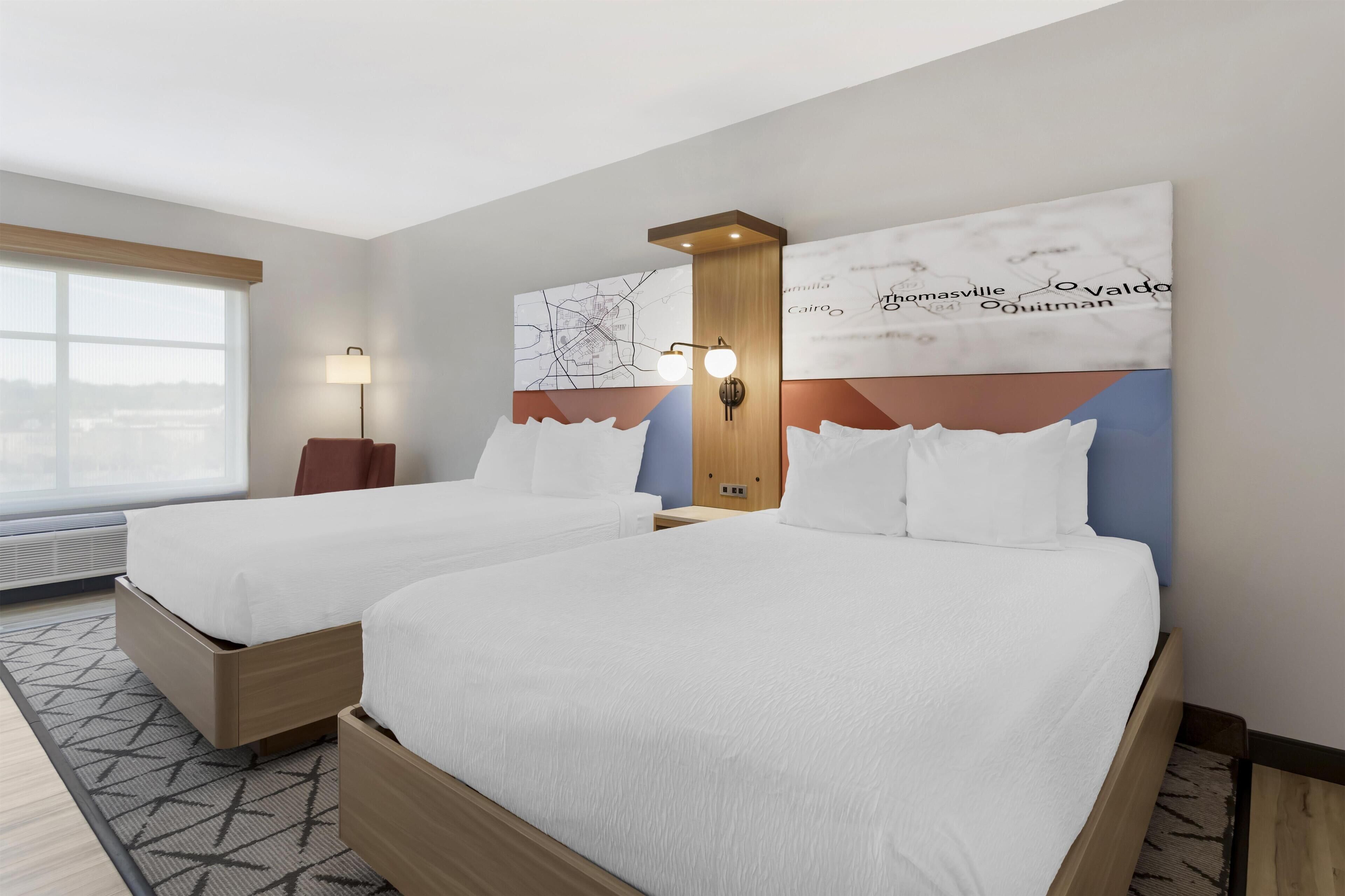 A hotel room with two beds, a large window, a map artwork on the wall, and a floor lamp.