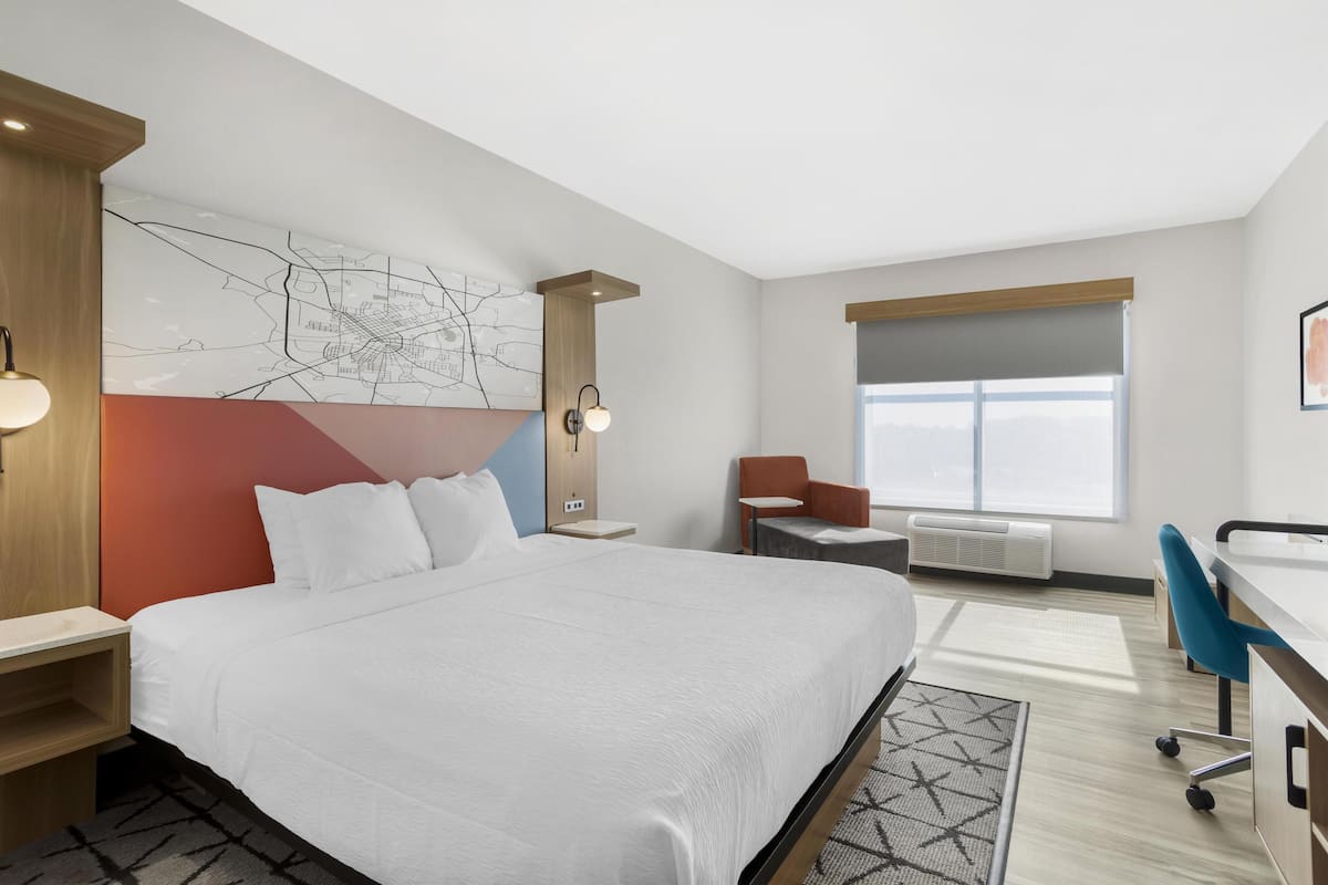 A modern hotel room with a large bed, a desk, a chair, a map artwork on the wall, and a large window.