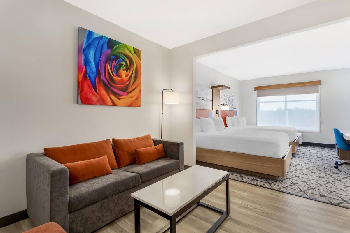 A modern hotel room with a gray sofa, a wooden bed, a large window, and a colorful abstract painting on the wall.