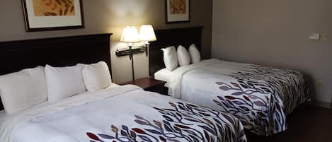 Dos camas con sábanas blancas y colchas de estampado de hojas, una mesita de noche con lámpara, y dos cuadros enmarcados en la pared.