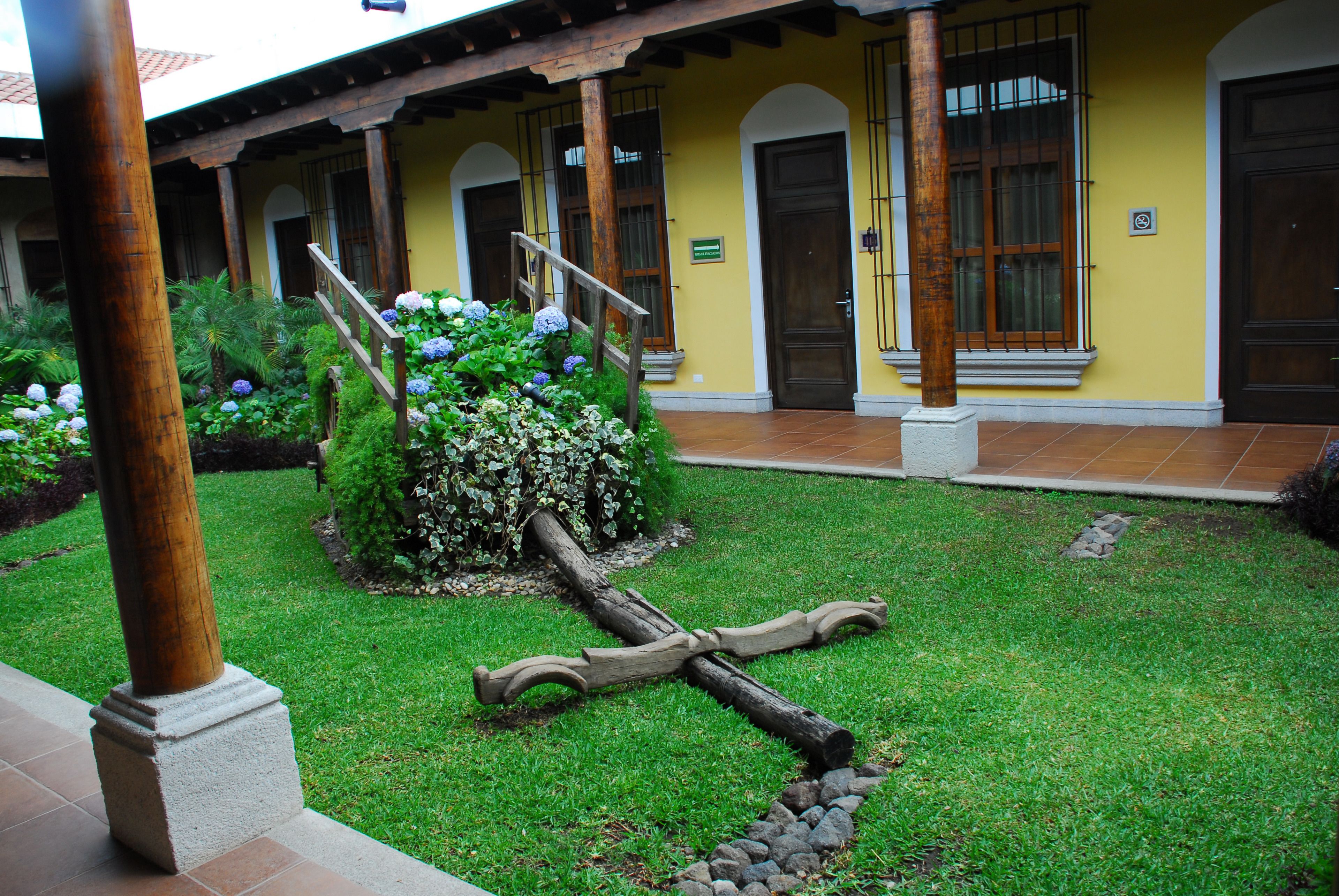 Um pátio com uma cruz de madeira, um pequeno jardim de hortênsias e um edifício amarelo com portas em arco.