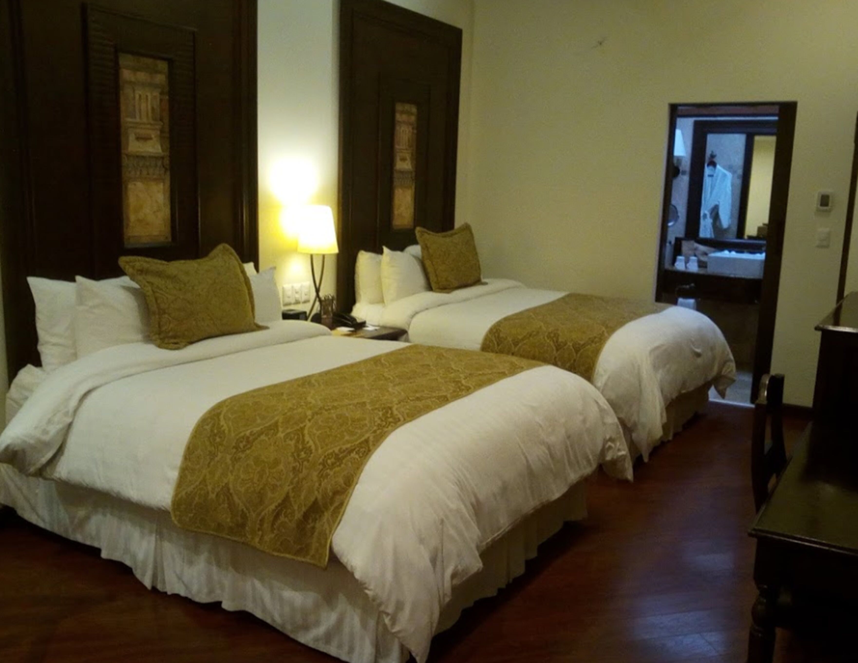 Ein Hotelzimmer mit zwei Betten, jeweils mit weißer Bettwäsche und einer braunen Tagesdecke. Dazwischen befindet sich ein Nachttisch mit einer Lampe, und rechts eine Holzkombination.