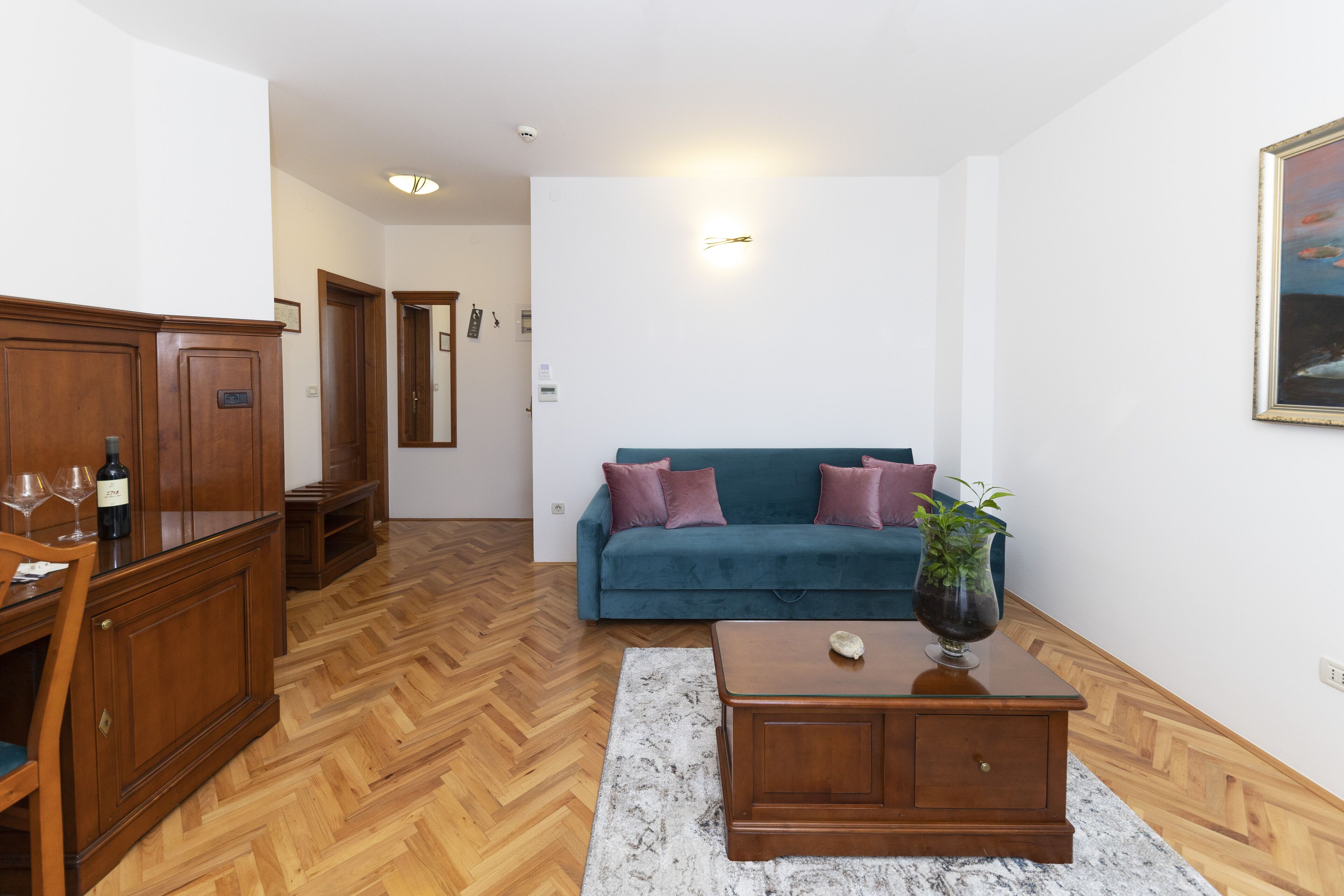 A living room with a wooden herringbone floor, a teal sofa with purple cushions, a wooden coffee table, and a cabinet with wine glasses.