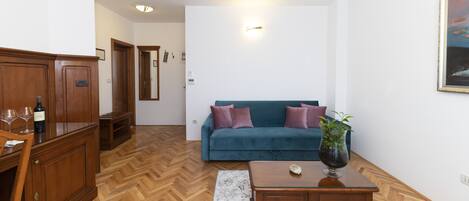 A living room with a wooden herringbone floor, a teal sofa with purple cushions, a wooden coffee table, and a cabinet with wine glasses.