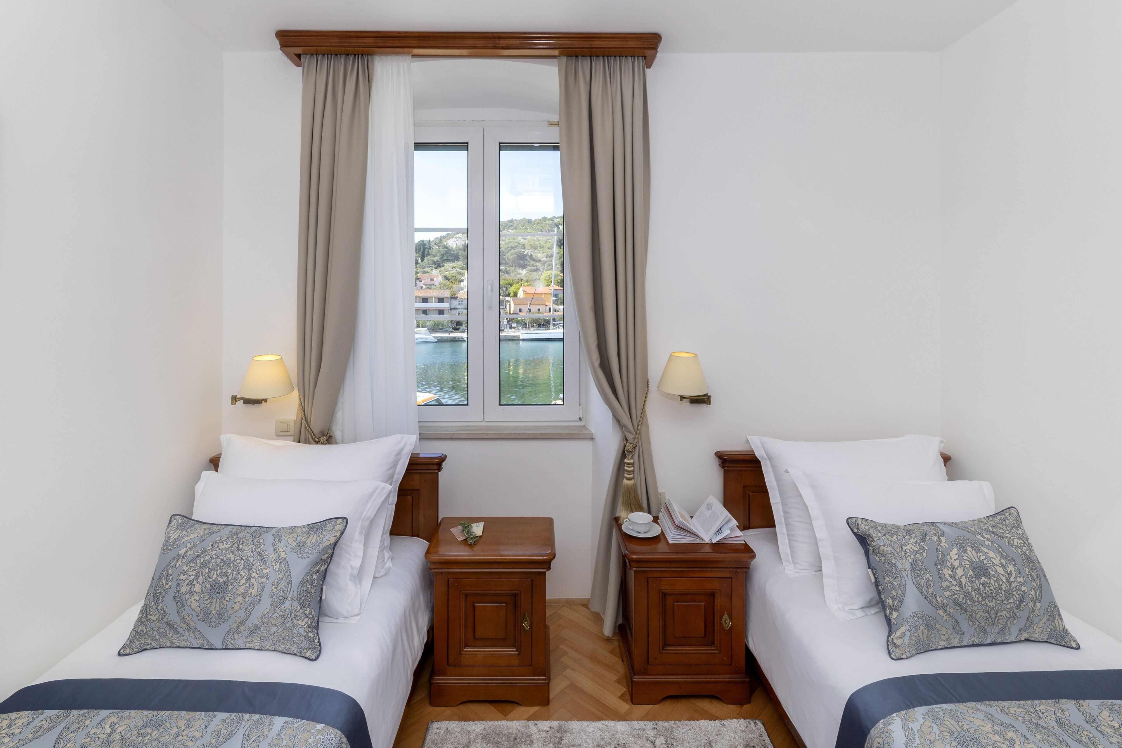 Quarto de hotel com duas camas, uma janela com cortinas, luminárias de cabeceira e vista para a água e edifícios.