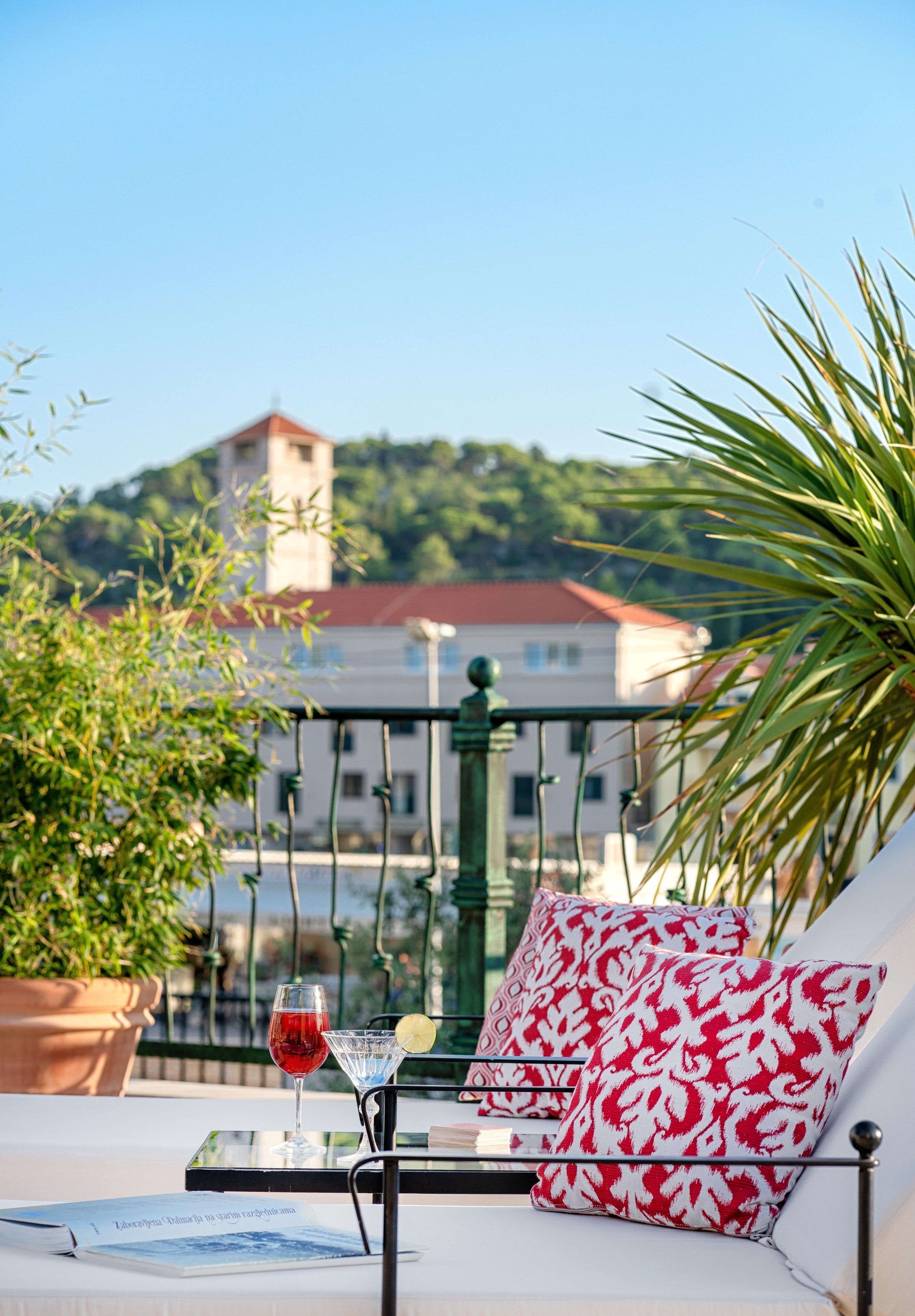 A balcony with a white sofa, red and white patterned pillows, a glass of red wine, and a lemon wedge. In the background, there is a building with a red roof and a tower.