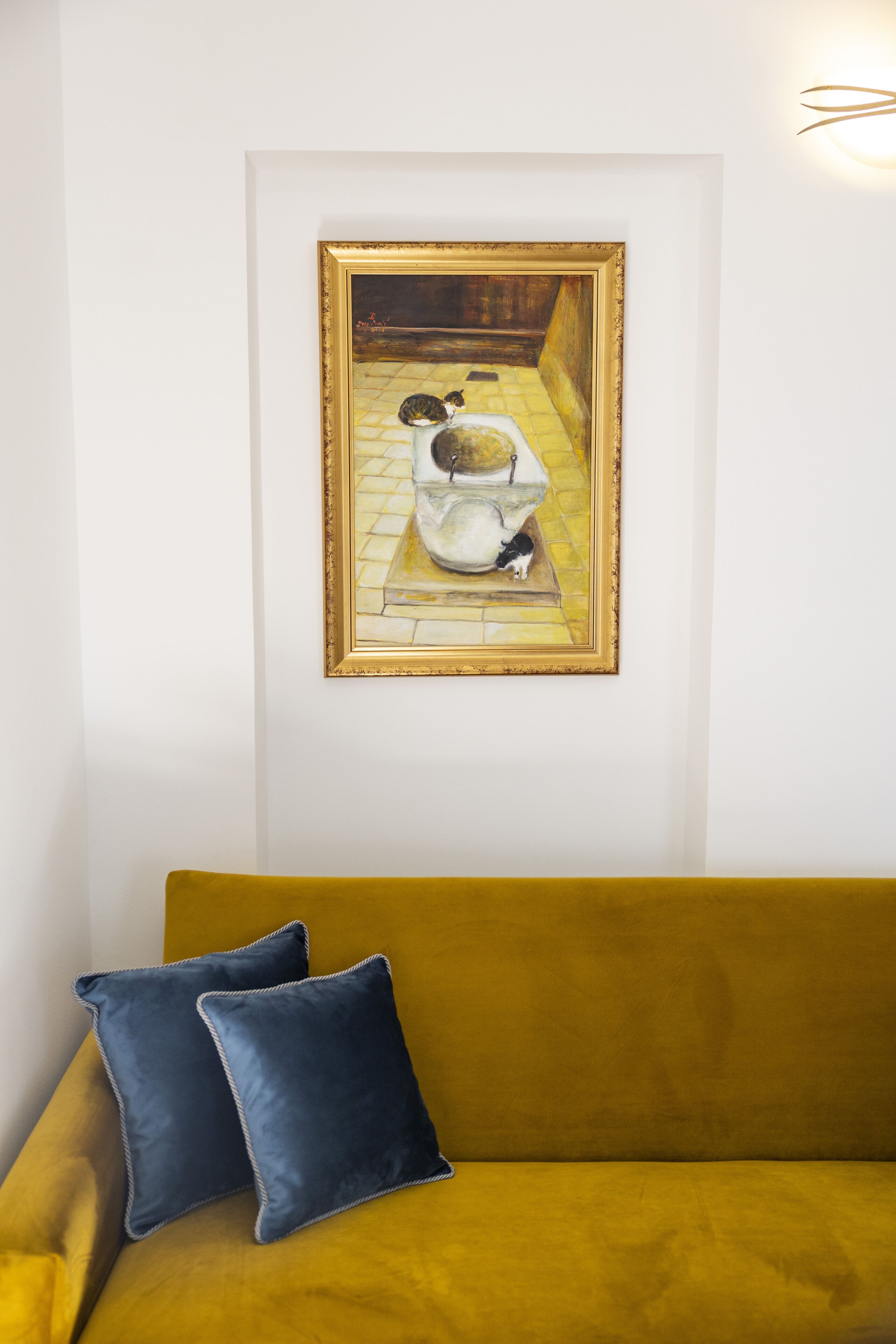 A room with a mustard yellow sofa and a framed painting of a bathroom scene.
