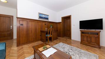 A room with wooden flooring, a dining table with chairs, a television on a wooden cabinet, and a painting on the wall.