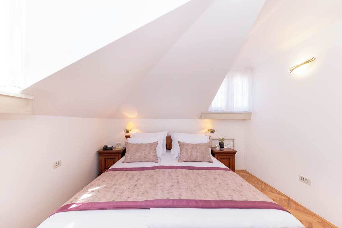 Un dormitorio con techo inclinado, una cama grande con colcha estampada, dos mesitas de noche con lámparas y una ventana pequeña.