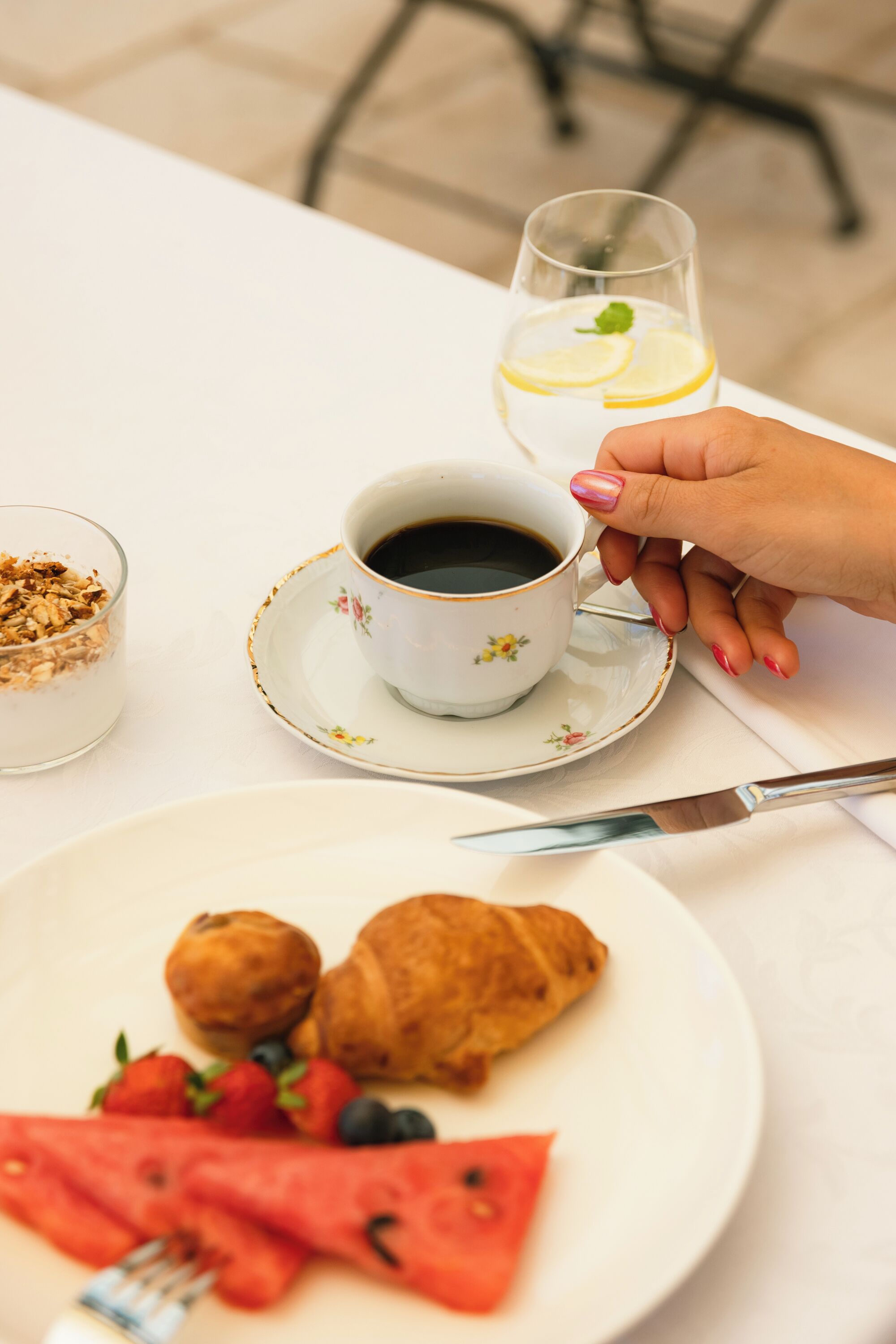 Un desayuno con una taza de café, un vaso de agua con rodajas de limón, un plato con croissants, fresas, arándanos y sandía.