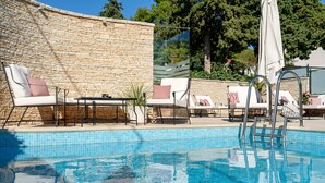 Piscina con sillas de descanso y una mesa, rodeada por un muro de piedra y árboles.