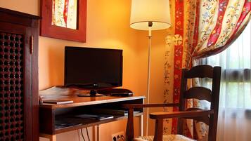 Habitación de hotel con escritorio de madera, televisor, lámpara, una silla y ventana con cortinas florales.