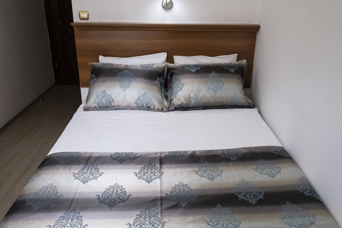 A neatly made bed with a patterned bedspread and matching pillows, a wooden headboard, and a wall-mounted light fixture.