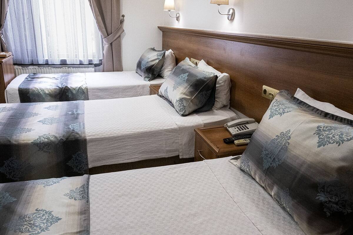 A hotel room with two beds, each with a white bedspread and matching pillows. There is a wooden headboard, a bedside table with a telephone and remote controls, and a window with curtains.