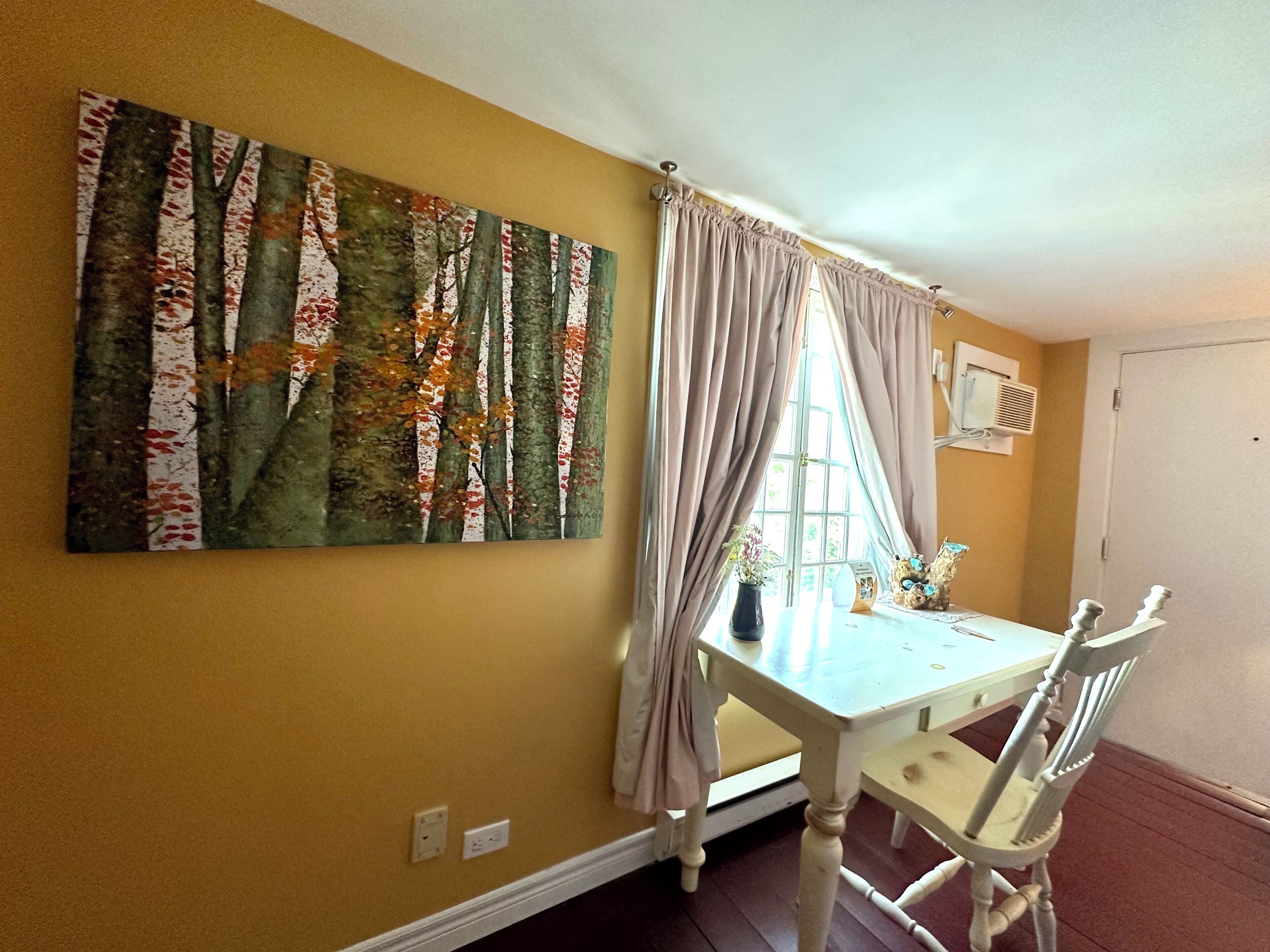 A dining area with a table and chairs, a window with curtains, and a painting on the wall.