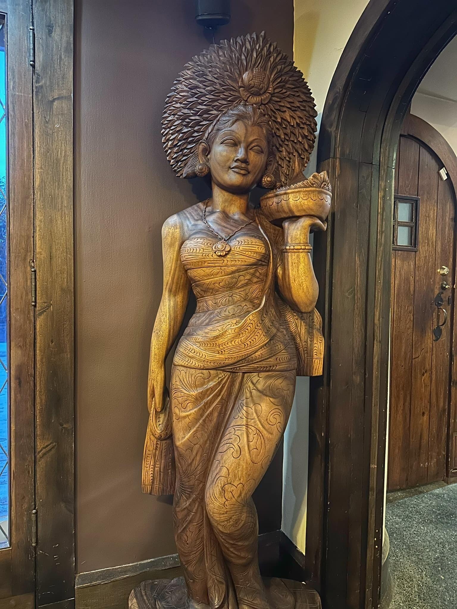 Una estatua de madera de una mujer con tallas detalladas, ubicada en un umbral con marco de madera y un ventanal pequeño.