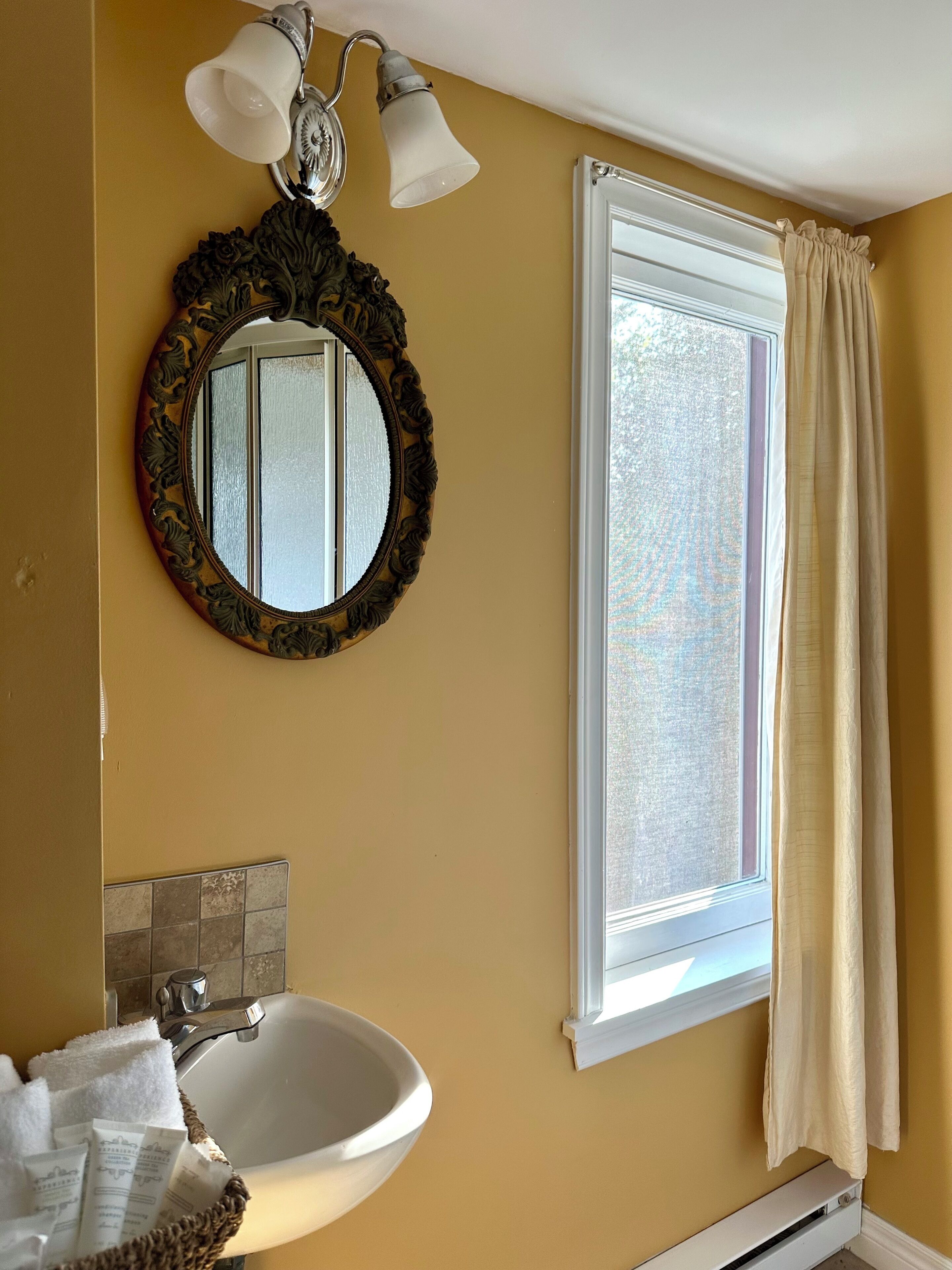 Een badkamer met een wit wastafelmeubel, een ronde spiegel met een sierlijk lijstwerk en een raam met een witte gordijn.