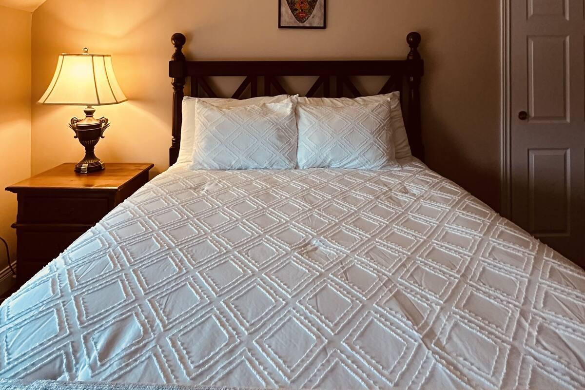 Una cama bien hecha con cabecera acolchada, una lámpara de noche y una obra de arte enmarcada en la pared.