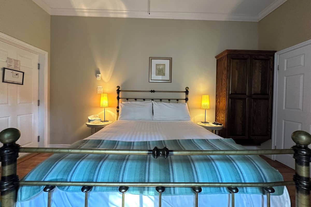 Un dormitorio ordenado con una cama centrada entre dos mesitas de noche, cada una con una lámpara. Un armario de madera está a la derecha, y un cuadro enmarcado cuelga en la pared sobre la cama.