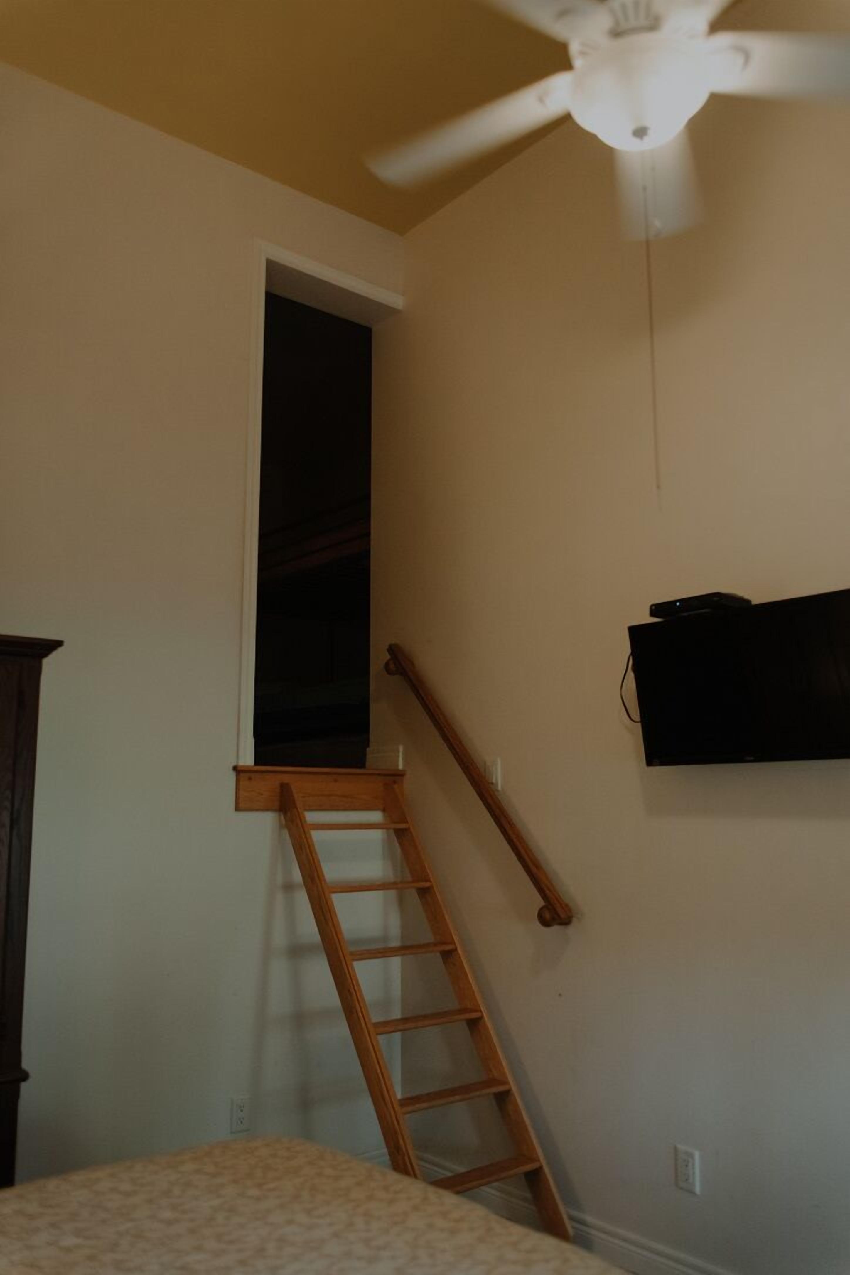 A room with a ceiling fan, a wooden ladder, a television mounted on the wall, and a window with a dark interior.