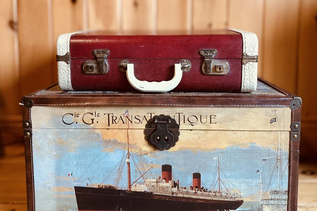 Una maleta antigua con asa y cierres metálicos sobre una caja de madera con una ilustración vintage de un barco y el texto "C.G. TRANSPORT UNIQUE".
