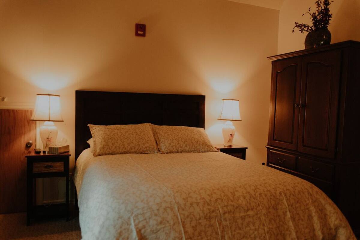 Una cama bien tendida con un cabecero oscuro, dos mesitas de noche con lámparas y un armario de madera.