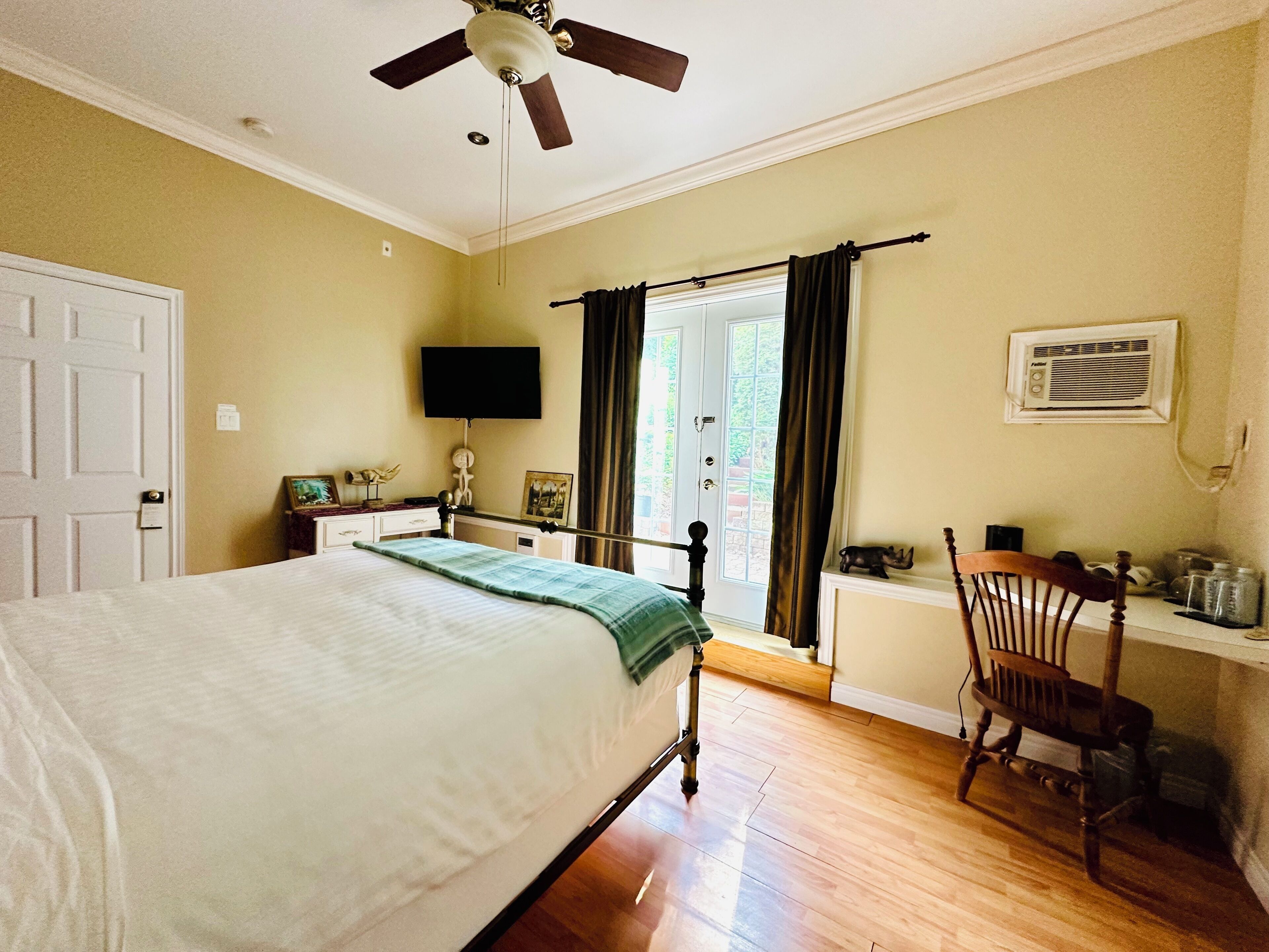 Un dormitorio con cama, ventilador de techo, televisor, ventana con cortinas, una silla y un escritorio con lámpara.