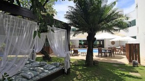 Un espace aménagé au bord de la piscine, comprenant un coin salon couvert, un palmier et du mobilier d’extérieur.