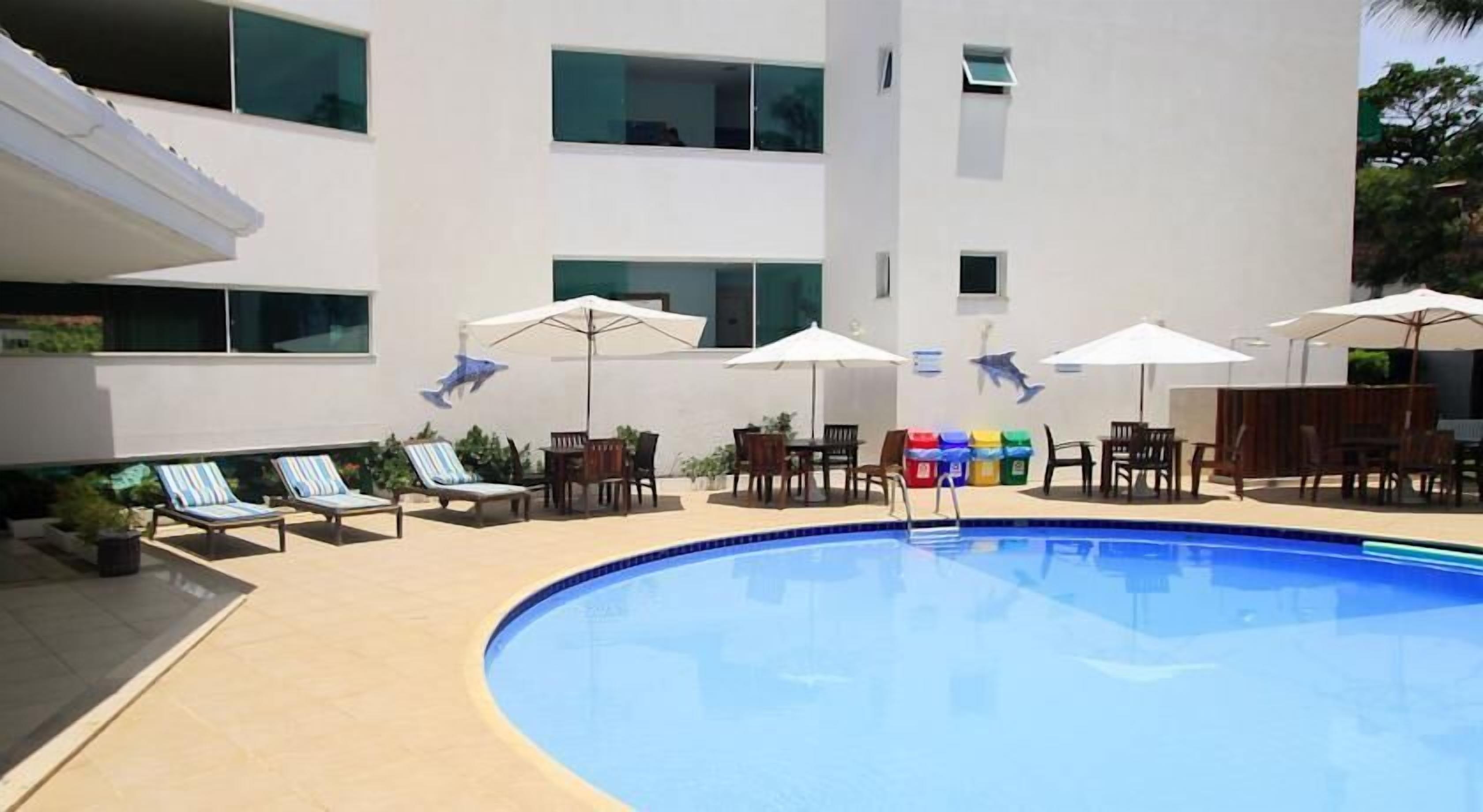 A swimming pool area with lounge chairs and umbrellas, surrounded by a building with multiple windows.