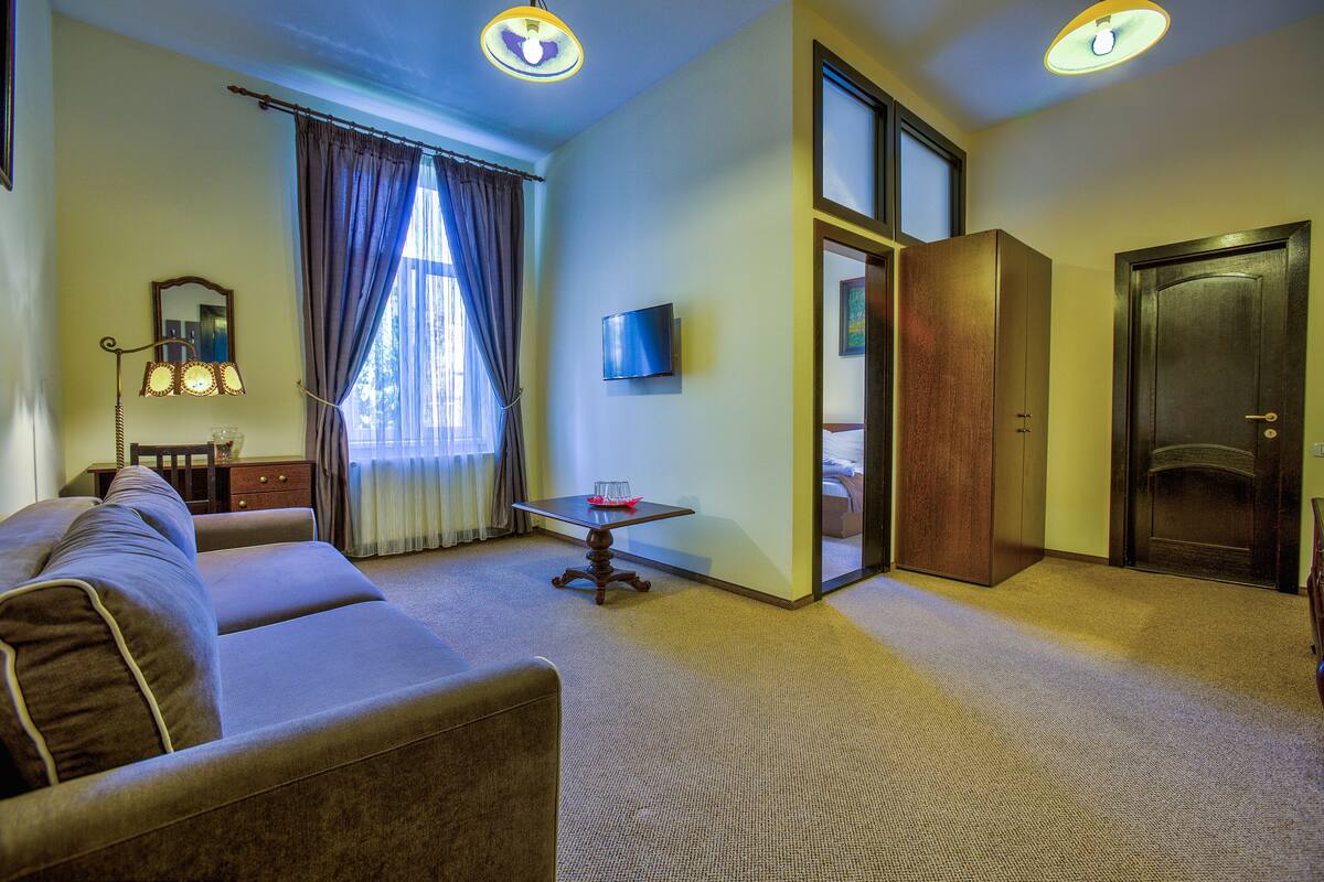 Quarto de hotel com sofá, mesa pequena, televisão, porta e janela com cortinas.