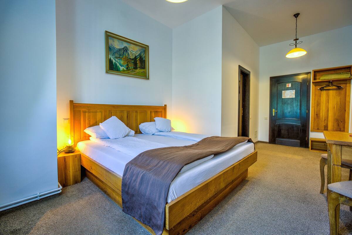 Quarto com cama de madeira, mesa de cabeceira com abajur, quadro na parede e porta que dá para outro cômodo.